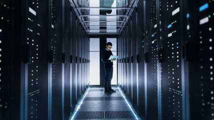 Male IT Engineer Works on a Laptop at the end of a Corridor in a Big Data Center. Rows of Rack...