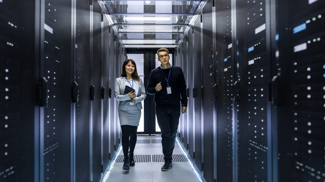 Caucasian Male and Asian Female IT Technicians Walking through Corridor of Data Center with Rows of Rack Servers. They Have Discussion, She Holds Tablet Computer. - Powered by Adobe