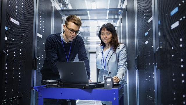 Caucasian Male and Asian Female IT Technicians Working with Computer Crash Cart in Big Data Center full of Rack Servers.