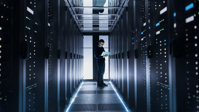 Male IT Engineer Works on a Laptop at the end of a Corridor in a Big Data Center. Rows of Rack Servers are Seen.