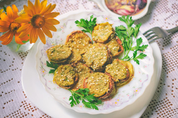 Rustic dinner with fried zucchini slices with nut sauce and greens. A white plate with food stands on a lacy rustic tablecloth, a bouquet with flowers in the background. Selective focus