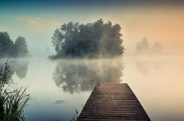 Morning misty landscape on the lake. Wooden pier and island with trees on the lake.