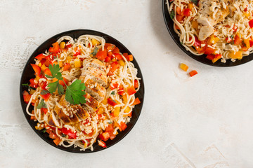 Spaghetti salad with fresh vegetables and baked chicken breast. Served on the black plates on the gray concrete table, top view.