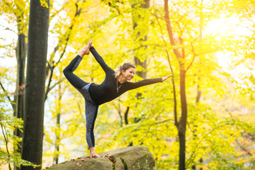 Sporty happy woman is practicing yoga and doing asana Narajasana in the autumn forest on the big boulder