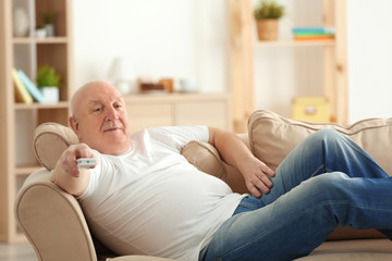 Fat senior man watching TV while lying on sofa at home. Sedentary lifestyle concept