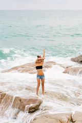 Pretty sporty girl in jeans short shorts, red headband and black bra stand on big stone on the beach during sea ocean storm. Big waves behind her. Concept of danger, sad, bad emotions.