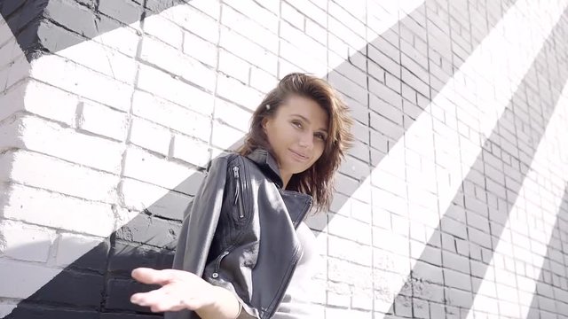 fun girl middle aged standing at the wall of brick with a striped color. On the shoulders of the leather jacket. luring gesture to follow her