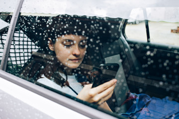 Bored and lonely girl sits inside the car while it rains outside, waits on her taxi or public transport, scrolls through application of social media on smartphone