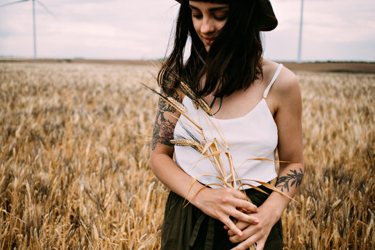 Femenine lady in thin silk white top and khaki panama hat and authentic hipster arm tattoos smiles to camera in middle of wheat field