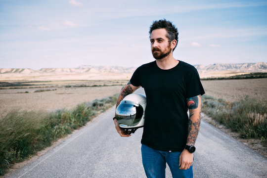 Brutal man with beard and arm tattoos poses with vintage retro helmet in blank mockup black tshirt in middle of american highway