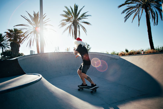 Funny cute man dressed like santa claus finally has summer vacation holidays, rides skateboard inside pool with amazing sun light leaks and palm trees