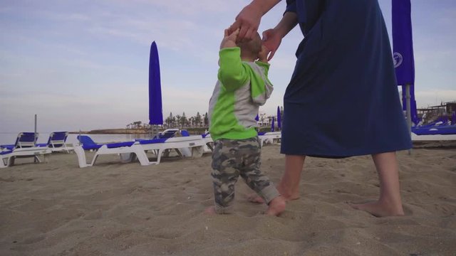 Learning to walk. Cute baby and his mother on a beach at evening time. Camera follows and moves around the family
