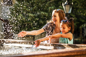 Mother and her daughter playing in the city square fountain. They sprayed with water. Refreshing on...