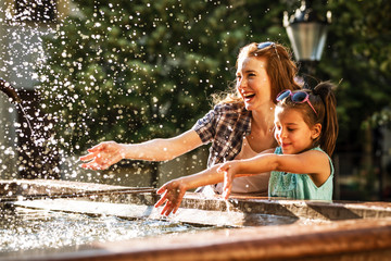 Mother and her daughter playing in the city square fountain. They sprayed with water. Refreshing on a hot summer day.	