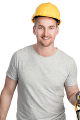 Smiling man in a grey shirt and green pants with a safety helmet standing in front of a white background holding a hammer and a lamp. Looking at camera.