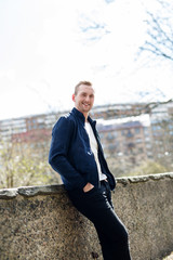 Fototapeta premium Handsome blonde man wearing a blue jacket and white tshirt leaning against a wall outdoors on a sunny day, trendy and fashionable.