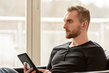 Pensive man wearing a black shirt and jeans sitting down indoors in a lounge chair holding a...
