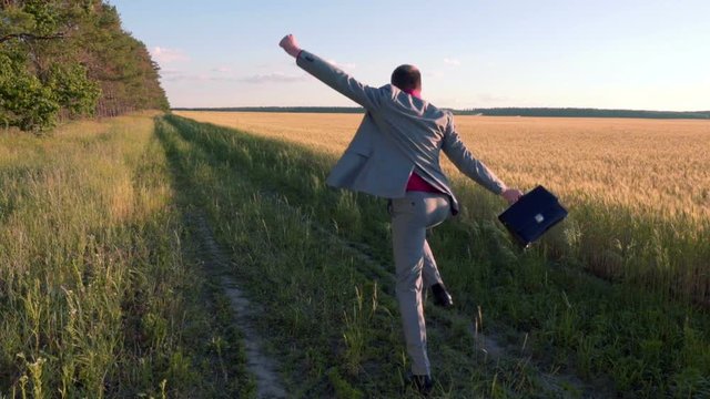 Rear view of a happy crazy businessman in a suit and with a leather briefcase runs along the wheat field waving his arms, slow motion