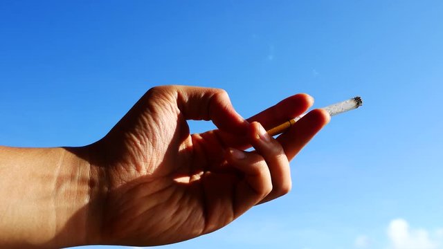 A human hand holding a smoking cigarette with blue sky background

