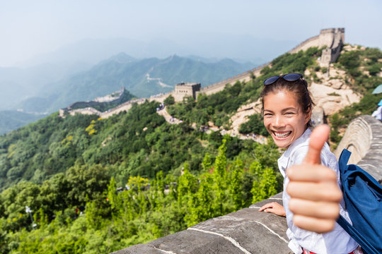 Happy tourist girl at Great Wall of china doing thumbs up hand sign having fun at famous Badaling ruins during travel holidays at Chinese destination. Asia vacation.