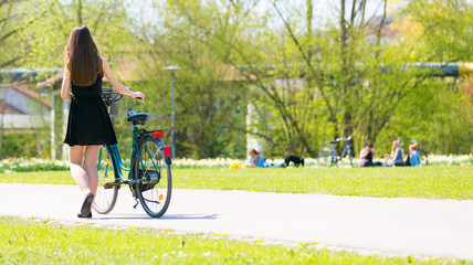 Obraz premium Back view of girl on bicycle wearing on black short dress. Young Woman riding along road on green spring outdoor Park. Sporty young girl riding a bicycle on a sunny morning, view from the back