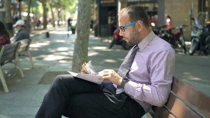 Young businessman eating sandwich working with documents on bench in city
 - Powered by Adobe