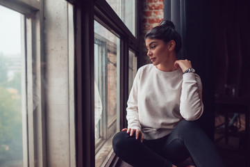 Attractive woman in gray sweatshirt sits on window sill and looks out the window. Mock-up.