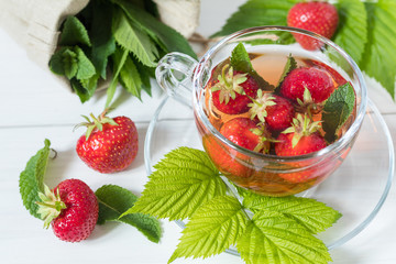 Glass cup of summer tea with fresh strawberry. Green leaves. Fresh mint. White wooden table. Shallow depth of field.