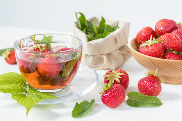 Glass cup of summer tea with fresh strawberry. Green leaves. Fresh mint. White wooden table. Shallow depth of field.