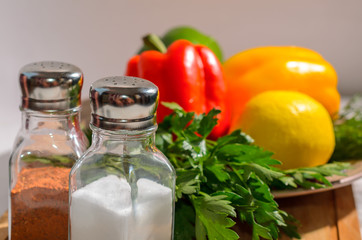 Salt shaker and pepper with fresh bright vegetables: Bulgarian pepper, herbs, lemon, on a ceramic plate, on a wooden cutting board
