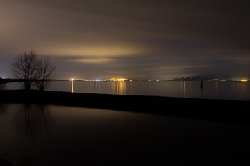 A nocturnal view of a lake, with perfectly still water, moving clouds, trees silhouettes and distant city lights