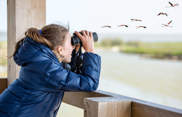 Girl with a winter jacket watching through binoculars from a wooden balcony - obrazy, fototapety, plakaty