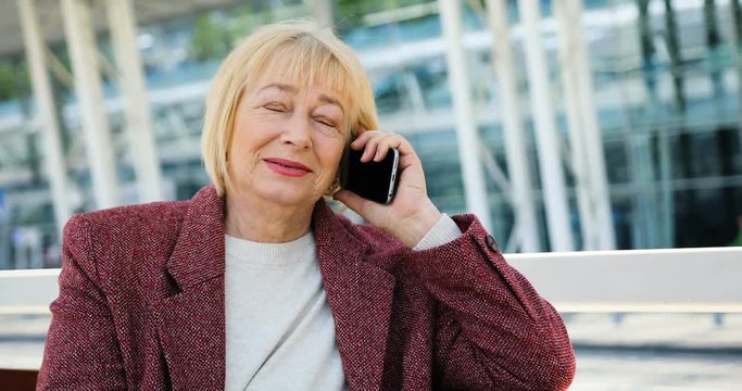 Beautiful older woman speaks on the phone outdoors in the sunlight near the business center