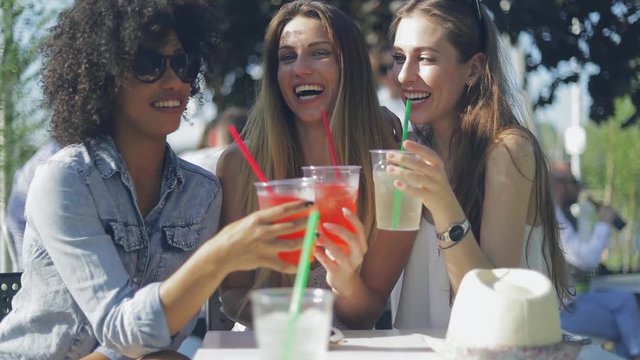 Young happy women wearing summer outfits clinking glasses with drinks while sitting at table in outside cafe and enjoying time together