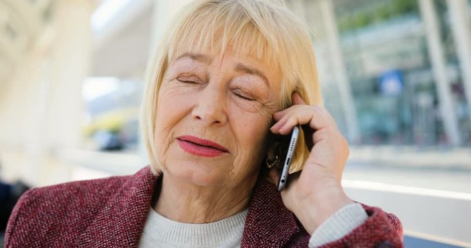 Beautiful older woman speaks on the phone outdoors in the sunlight near the business center