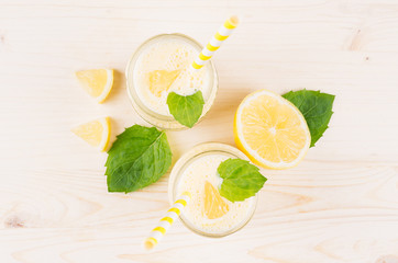 Freshly blended yellow lemon smoothie in glass jars with straw, mint leaf, cut lemon,  top view, close up. White wooden board background.