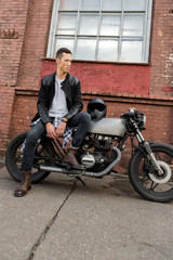 Plakat Handsome rider biker man in black leather jacket, jeans and boots sit on classic style cafe racer motorbike. Bike custom made in vintage garage. Brutal fun urban lifestyle. Outdoor portrait.