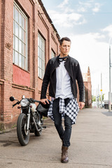 Handsome rider biker guy in black leather jacket and helmet walk away from classic style cafe racer motorcycle. Bike custom made in vintage garage. Brutal fun urban lifestyle. Outdoor portrait.