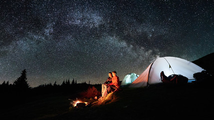 Night camping in the mountains. Man and woman tourists sitting at a campfire near two illuminated...