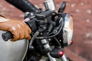 Close up of a hipster biker man hand in leather glove hold throttle control of classic style cafe racer motorcycle. Bike custom made in vintage garage. Brutal fun urban lifestyle. Outdoor portrait.