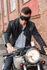 Naklejka na ściany i meble Handsome rider biker man in black leather jacket and protection googles sit on classic style cafe racer motorcycle. Bike custom made in vintage garage. Brutal fun urban lifestyle. Outdoor portrait.