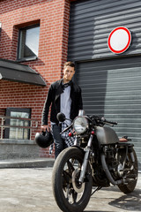 Handsome young rider guy in black leather biker jacket go to his classic style cafe racer motorcycle industrial gates as background. Bike custom made in vintage garage. Brutal fun urban lifestyle.
