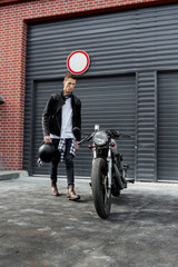 Handsome young rider man in black biker jacket and boots go to his classic style cafe racer motorcycle industrial gates as background. Bike custom made in vintage garage. Brutal fun urban lifestyle.