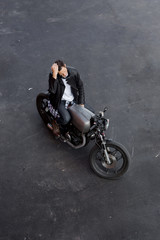 Plakat Top view of a handsome rider man in black biker jacket and checkered shirt correct hes hair sit on classic style cafe racer motorcycle. Bike custom made in vintage garage. Brutal fun urban lifestyle.