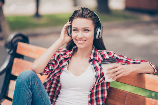 Nice spring weekend! Close up of a dreamy young lady, listening to music outside, sitting on the wooden bench in the park, wearing cozy outfit, modern headphones, with tea
