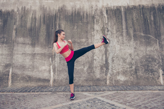 Side profile photo of attractive brunette, kickboxing high kick exercise with her foot outdoors near concrete wall, wearing fashionable pink and black sport wear - Powered by Adobe