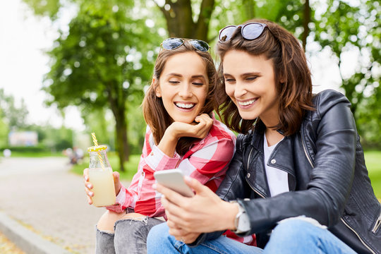 Two Female Friends Spending Time Together At Park Browsing Internet On The Phone