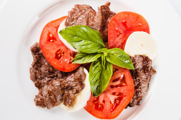 Meat salad. Fried beef with mozzarella, tomato and basil on a white plate on a light background (top view  close)