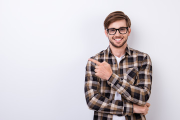 Smiling young nerdy bearded stylish student is standing on pure background in glasses and casual  outfit, pointing on the copyspace - obrazy, fototapety, plakaty