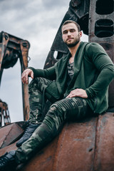 Close up fashion portrait of stylish handsome man sitting on rusty elements. Man in clothes khaki colors on city of urban background.